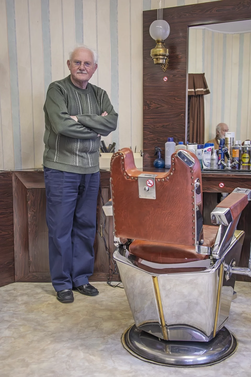 Portrait d'un barbier âgé, bras croisés, à côté d'un fauteuil de coiffeur ancien dans son salon