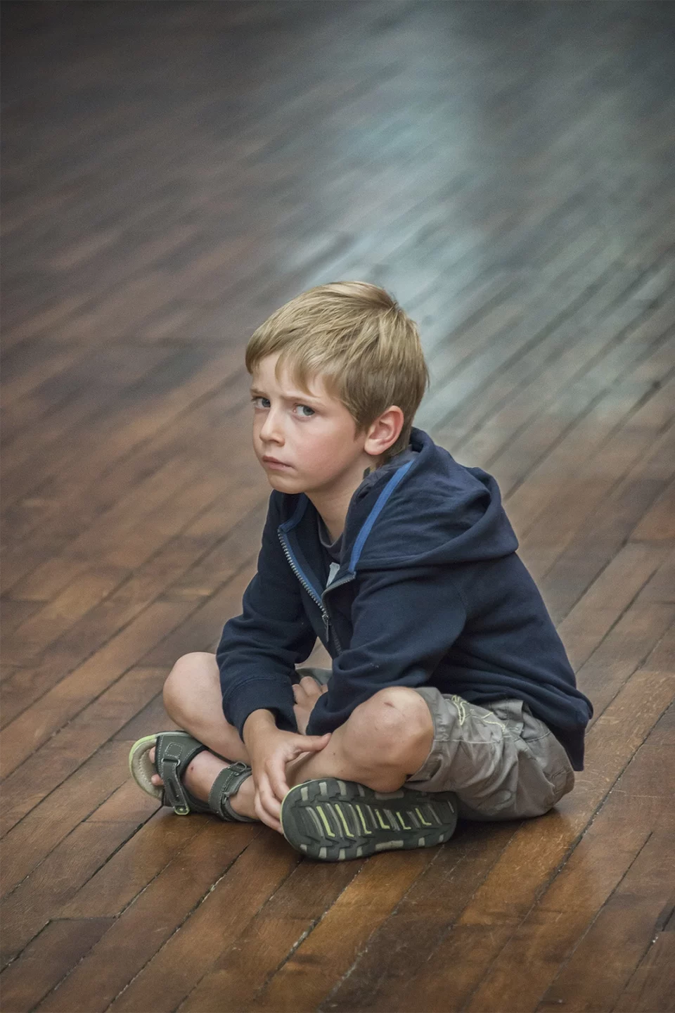 Portrait d'un petit garçon blond assis en tailleur sur un parquet, le regard tourné de côté
