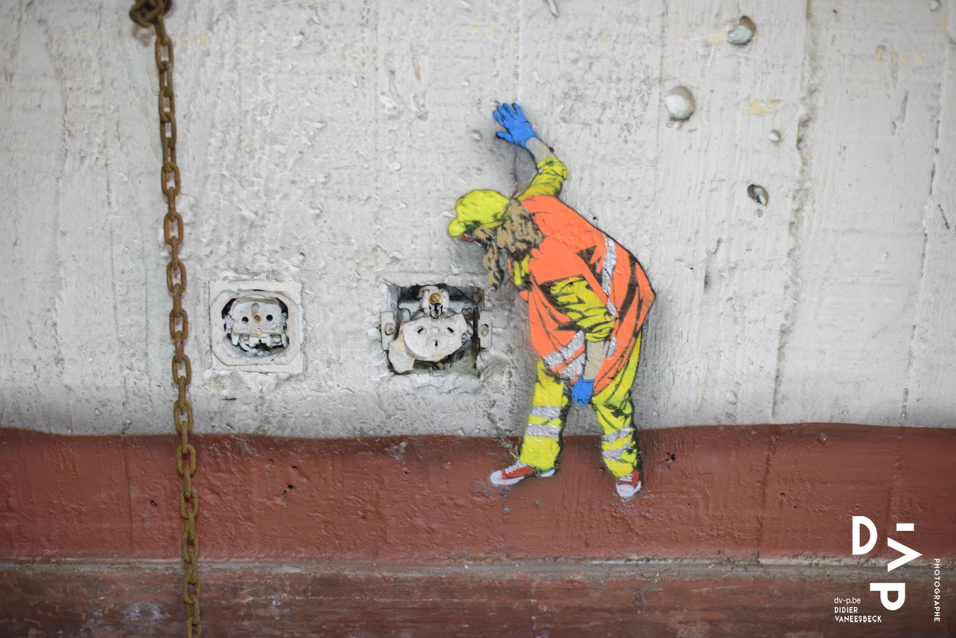 Personnage peint en orange et bleu sur un mur, chaîne suspendue – Strokar Inside, Bruxelles 2018